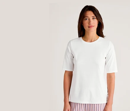 Frau mit dunklen Haaren trägt ein weißes CALIDA Kurzarm-Sleep-T-Shirt und eine rosa-weiß gestreifte Hose.