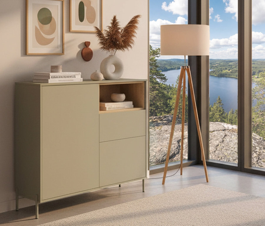 Ein salbeigrünes Highboard »Hygge« mit zwei Türen steht neben einer Stehlampe vor einem Fenster mit Seeblick.