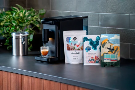 Espressomaschine befüllt Glas, daneben drei Kaffeepackungen Alltagsheld, Community Blend und Bergsonne. Dose links.