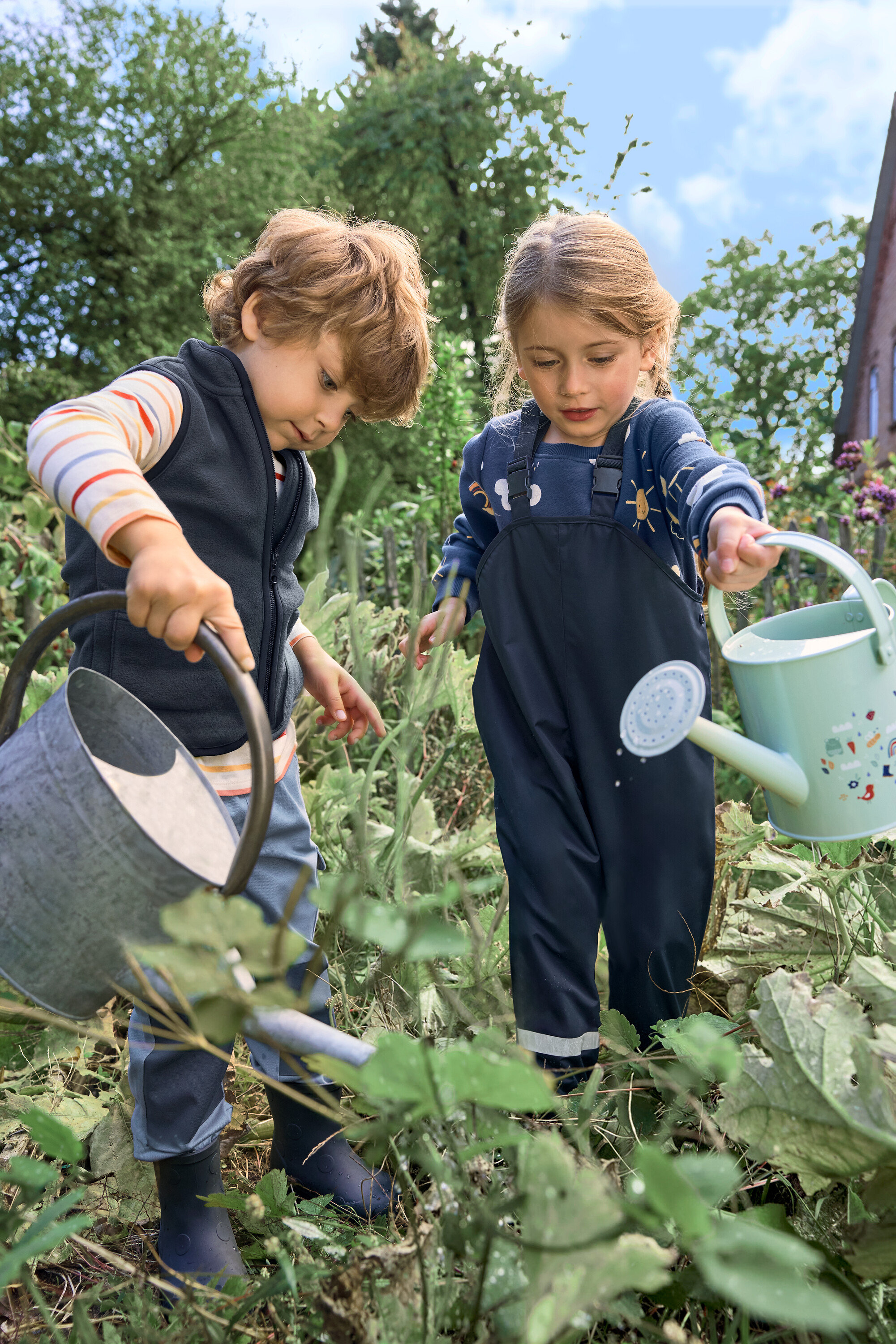 Zwei Kinder gießen Pflanzen im Garten. Ein Kind trägt dunkelblaue Kinder-Thermostiefel.