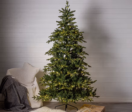 Ein hoher, grüner künstlicher Weihnachtsbaum mit warmweißen Lichtern steht auf dunklem Boden vor einer weißen Holzwand. Links daneben ein heller Sessel mit einer grauen Decke und einem cremefarbenen Kissen. Am Boden liegt ein weißer Schaffellteppich, unter dem Baum zwei braun verpackte Geschenke.