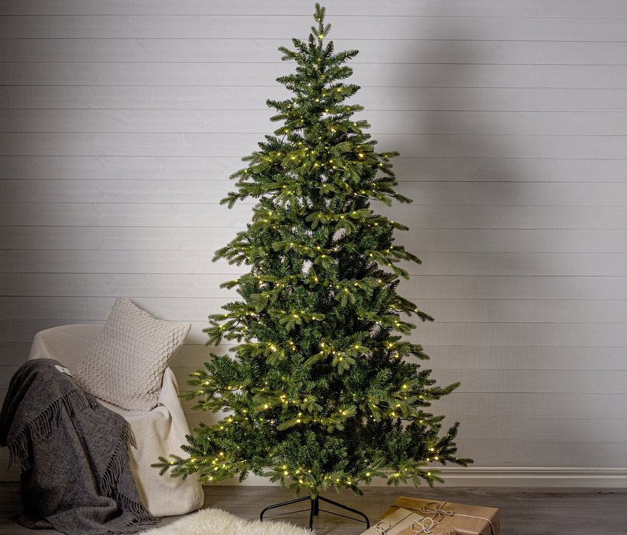 Ein hoher, grüner künstlicher Weihnachtsbaum mit warmweißen Lichtern steht auf dunklem Boden vor einer weißen Holzwand. Links daneben ein heller Sessel mit einer grauen Decke und einem cremefarbenen Kissen. Am Boden liegt ein weißer Schaffellteppich, unter dem Baum zwei braun verpackte Geschenke.