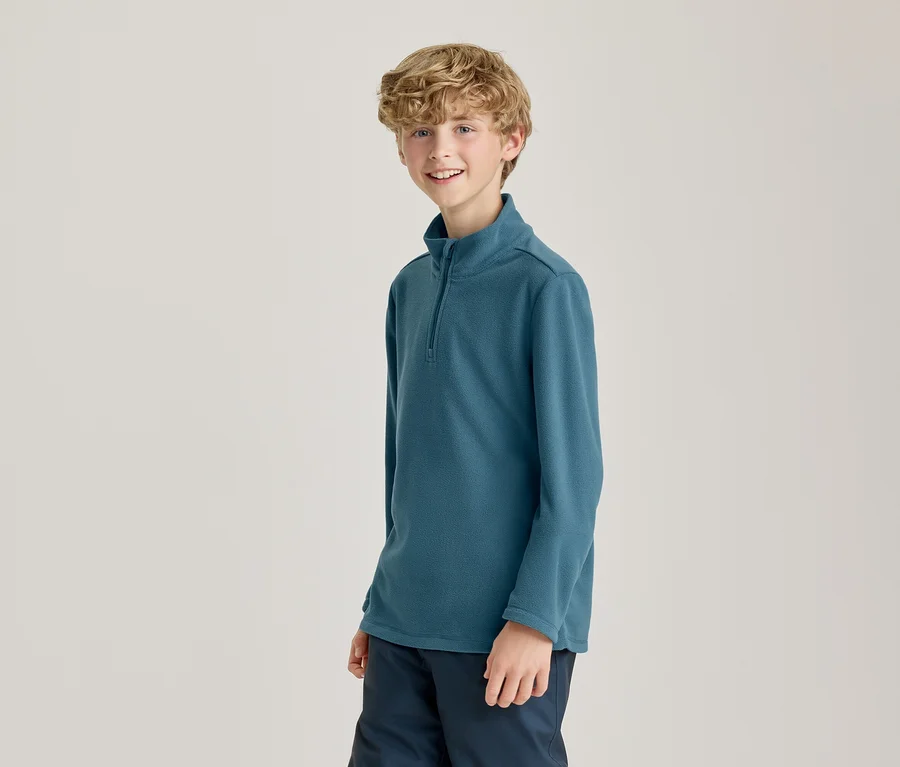 Ein lächelnder junger Junge mit lockigem blondem Haar trägt ein türkisfarbenes Langarmshirt mit Stehkragen und halbem Reißverschluss sowie eine dunkelblaue Hose vor hellem Hintergrund.