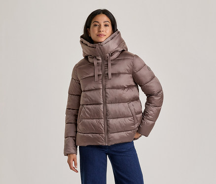Frau in einer braunen Steppjacke mit hohem Kragen und Kapuze sowie blauen Jeans, die mit den Händen in den Taschen steht.
