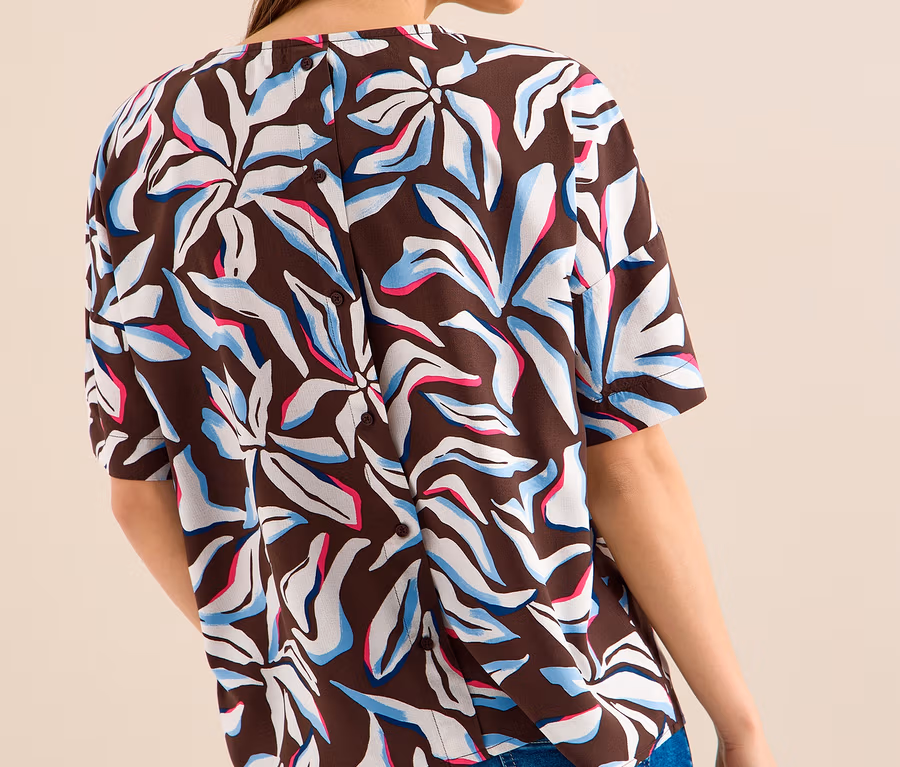 Frau von hinten, in einer braunen CECIL Kurzarmbluse mit weißen, hellblauen und pinken abstrakten Blumenmustern und Knöpfen am Rücken, dazu blaue Jeans.