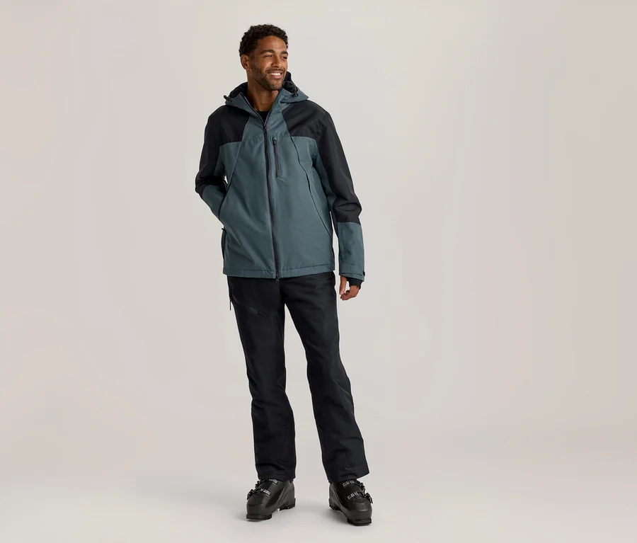 Mann steht lächelnd und blickt nach rechts, trägt eine türkis-schwarze Skijacke, schwarze Skihose und schwarze Skischuhe.