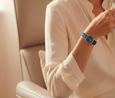 Arm einer Person mit cremefarbener Bluse, die eine Uhr mit blauem Zifferblatt und blauem Lederarmband trägt und eine Tasse hält.