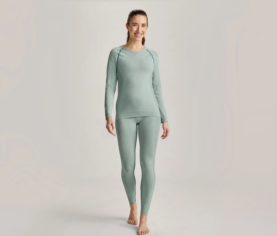 Lächelnde Frau steht in hellgrünem Langarmshirt und passender Leggings mit dunklen Nähten, barfuß.