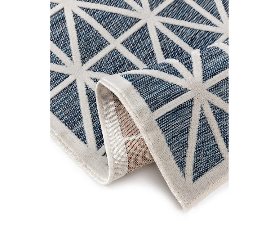Ein Teppich mit blauem, gewebtem Hintergrund und weißen geometrischen Mustern. Die weißen Linien bilden miteinander verbundene Dreiecke und sternähnliche Formen. Der Teppich ist teilweise aufgerollt und zeigt seine hellere Unterseite. Er hat einen weißen Rand.