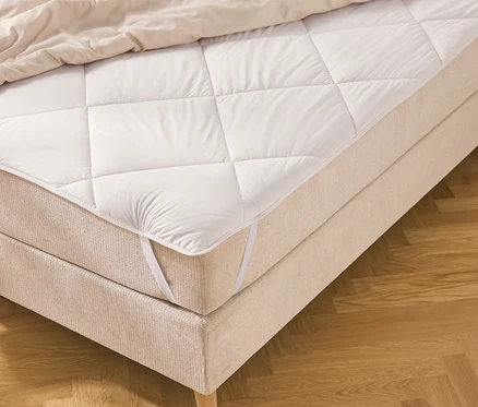 Ein Bett mit beigem Stoffbezug und einer weißen, gesteppten Matratzenauflage, die mit Gummibändern befestigt ist. Eine helle Bettdecke liegt am oberen Ende, das Bett steht auf einem Fischgrätparkettboden.