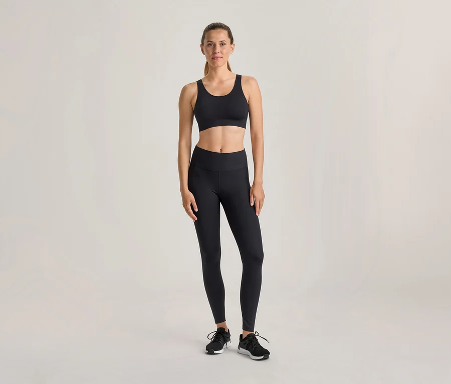 Eine Frau steht in einem schwarzen Sport-BH, schwarzen Leggings und schwarzen Sportschuhen vor einem hellen Hintergrund.