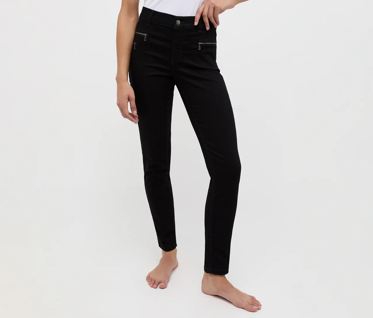Person trägt schwarze Skinny Jeans mit Reißverschlussdetails an den Vordertaschen, barfuß vor weißem Hintergrund stehend.