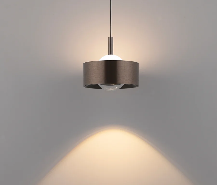 Eine moderne Pendelleuchte mit zylindrischem Schirm aus gebürstetem Metall hängt an einem schwarzen Kabel und wirft einen warmen, hellen Lichtbogen auf eine hellgraue Wand.