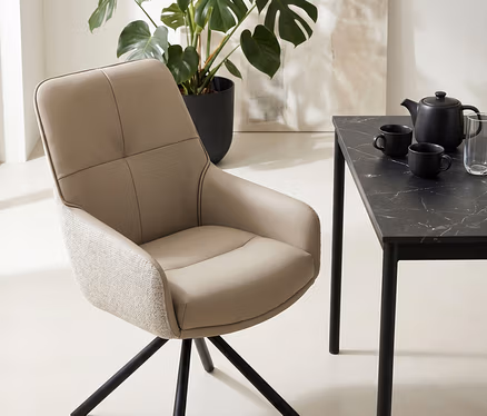 Ein beiger Esszimmerstuhl mit schwarzem Metallfuß steht neben einem schwarzen Tisch mit Marmoroptik, auf dem eine Teekanne, zwei Tassen und ein Glas stehen. Im Hintergrund sind eine große Monstera-Pflanze und ein abstraktes Gemälde zu sehen.