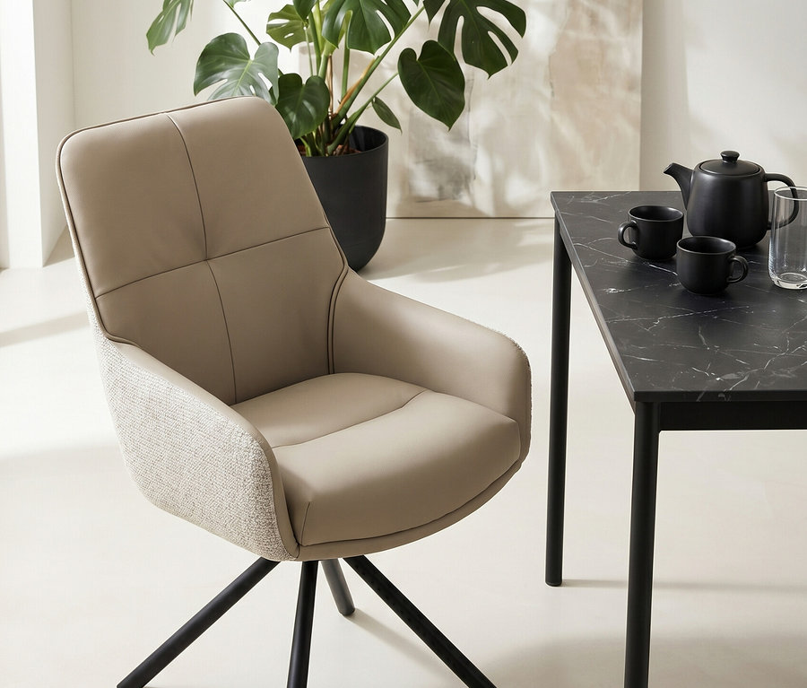 Ein beiger Esszimmerstuhl mit schwarzem Metallfuß steht neben einem schwarzen Tisch mit Marmoroptik, auf dem eine Teekanne, zwei Tassen und ein Glas stehen. Im Hintergrund sind eine große Monstera-Pflanze und ein abstraktes Gemälde zu sehen.