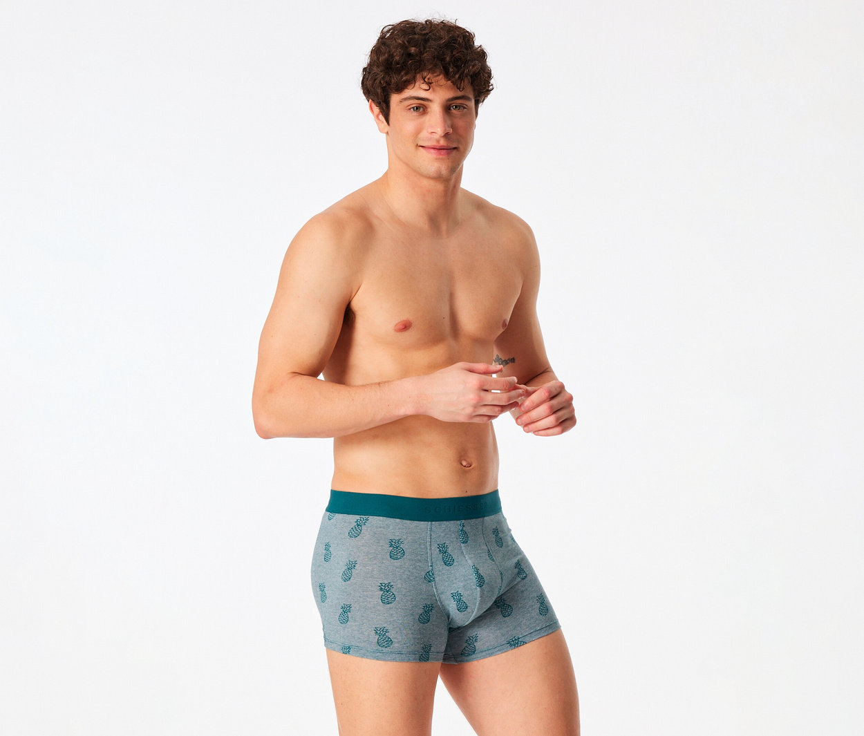 Ein lächelnder Mann mit lockigem Haar, der graue Boxershorts mit Ananasmuster und einem dunkelgrünen Bund trägt, steht vor einem weißen Hintergrund.