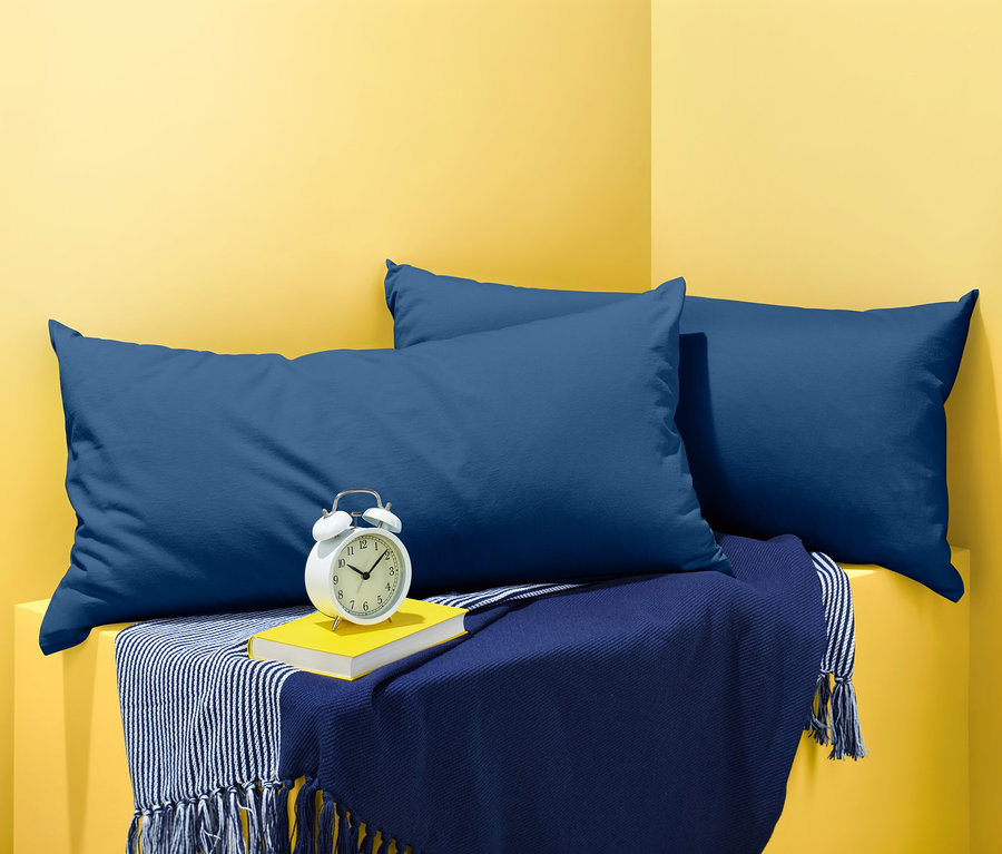 Zwei blaue Jersey-Kopfkissenbezüge liegen auf einer blauen Decke mit einem Buch und einem Wecker.