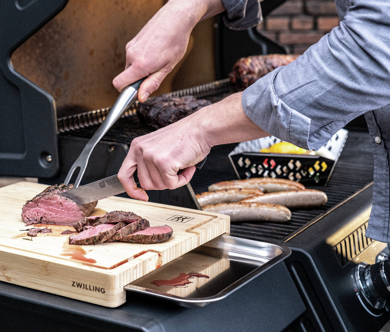 Eine Person schneidet einen Braten auf einem ZWILLING Schneidebrett mit Auffangschale, daneben Würste auf einem Grill.