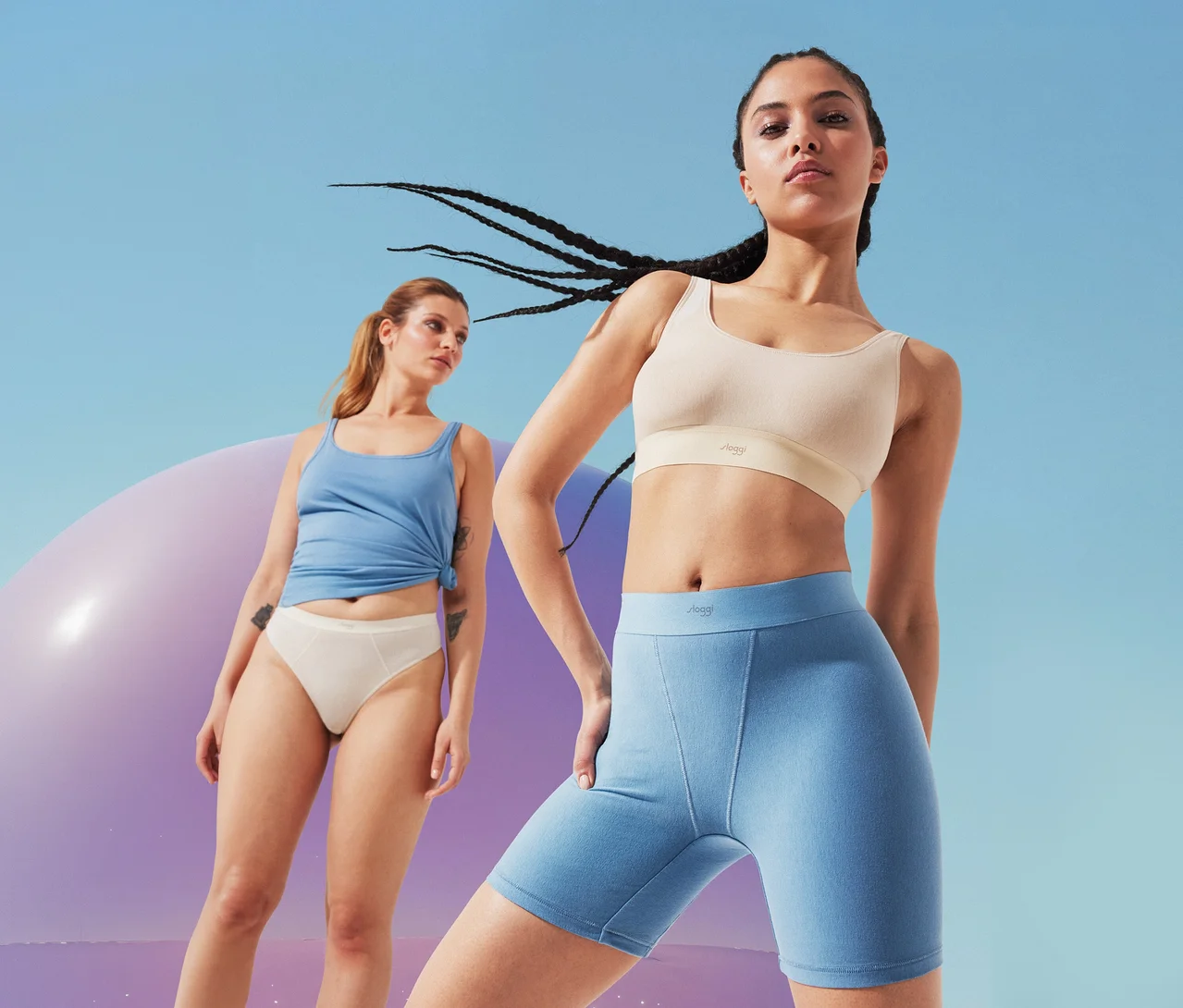 Zwei Frauen posieren vor blauem Himmel und lila Objekt. Eine Frau im Vordergrund trägt beigen Sport-BH und hellblaue Radlerhosen, die andere im Hintergrund hellblaues Top und beigen Slip.