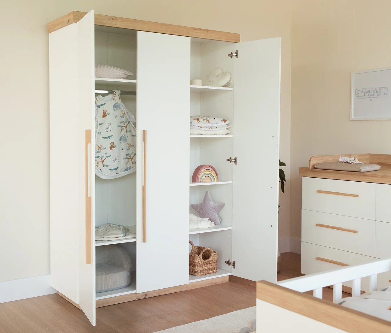 Ein offener weißer und hölzerner Kleiderschrank in einem Kinderzimmer, gefüllt mit einem Babyschlafsack mit Tiermuster, gefalteter Kleidung, einem grauen Babysitz, einem Regenbogenspielzeug und einem Sternkissen auf Regalen. Eine weiße Wickelkommode mit Holzplatte und ein Babybett sind ebenfalls sichtbar.