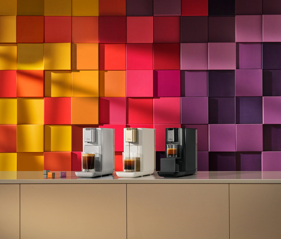 Drei Kaffeemaschinen in Hellgrau, Creme und Schwarz stehen auf einer beigen Theke. Die cremefarbene Maschine brüht Kaffee in ein Glas. Dahinter ist eine Wand aus quadratischen Fliesen mit einem Farbverlauf von Gelb und Orange links zu Rot, Pink und Lila rechts. Kaffeekapseln liegen ebenfalls auf der Theke.