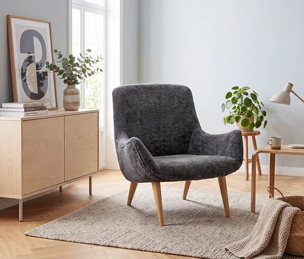 Ein dunkelgrauer Sessel auf einem hell gemusterten Teppich in einem hellen Wohnzimmer. Links ein Holzsideboard mit Kunst, Vase und Büchern, dahinter ein Fenster. Rechts ein Beistelltisch mit Lampe, Tasse, Pflanze und Korb mit Decke.