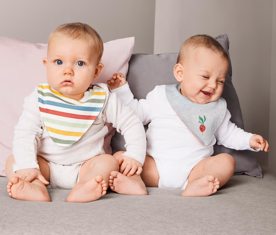 Zwei Babys sitzen nebeneinander auf einer grauen Decke und tragen 2 Dreieckstücher. Eines hat ein Tuch mit bunten Streifen, das andere mit einem Radieschenmuster.