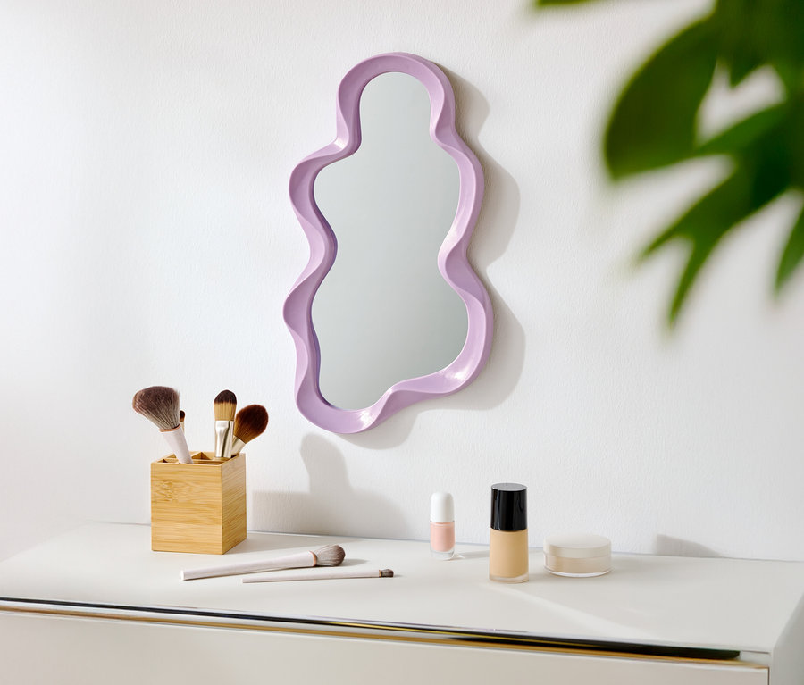 Ein lilafarbener, unregelmäßig geformter Spiegel hängt an einer weißen Wand über einem weißen Tisch mit Make-up-Pinseln und Kosmetika.