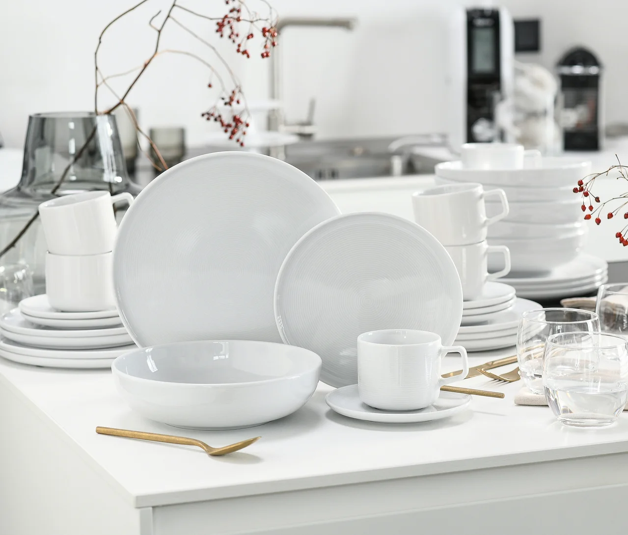 Ein weisses creaTable Service "Session White", 30-teilig, ist auf einer weissen Fläche arrangiert, mit Tellern, Schüsseln und Tassen mit Untertassen, teils gestapelt, neben goldenem Besteck und klaren Gläsern.