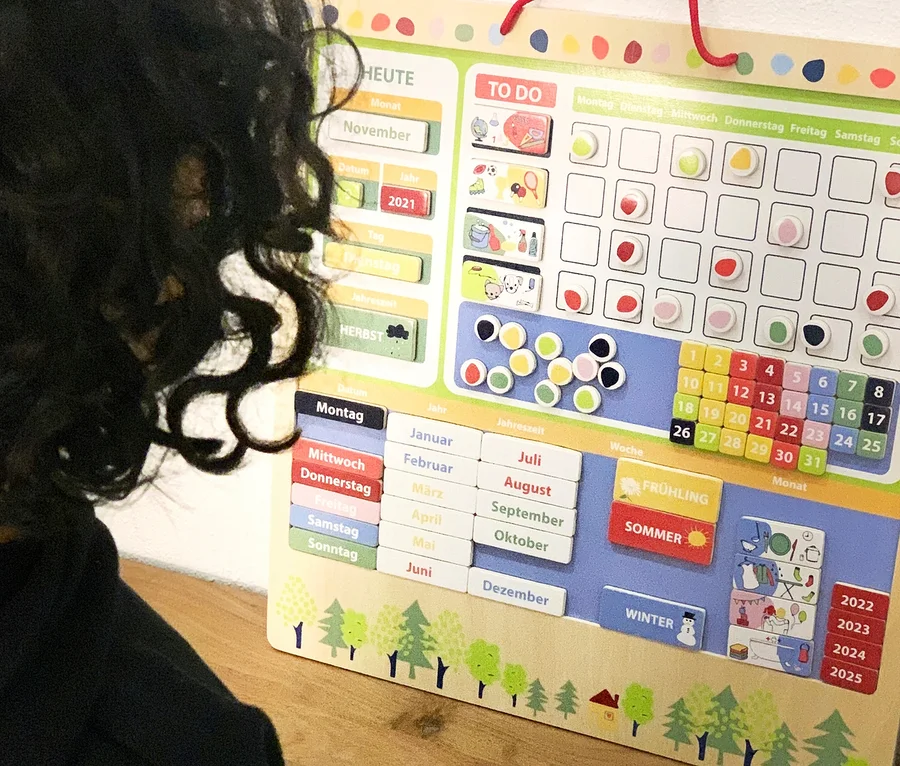 Kind hält eine Tasse vor einem magnetischen Wochenplaner »Educate« von small foot. Der Planer zeigt die Wochentage, Monate und Jahreszeiten.