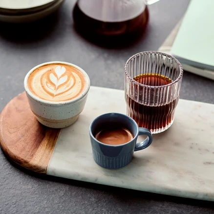 Auf einem Marmorbrett stehen ein Cappuccino, ein Espresso in einer blauen Tasse und ein Glas Kaffee.