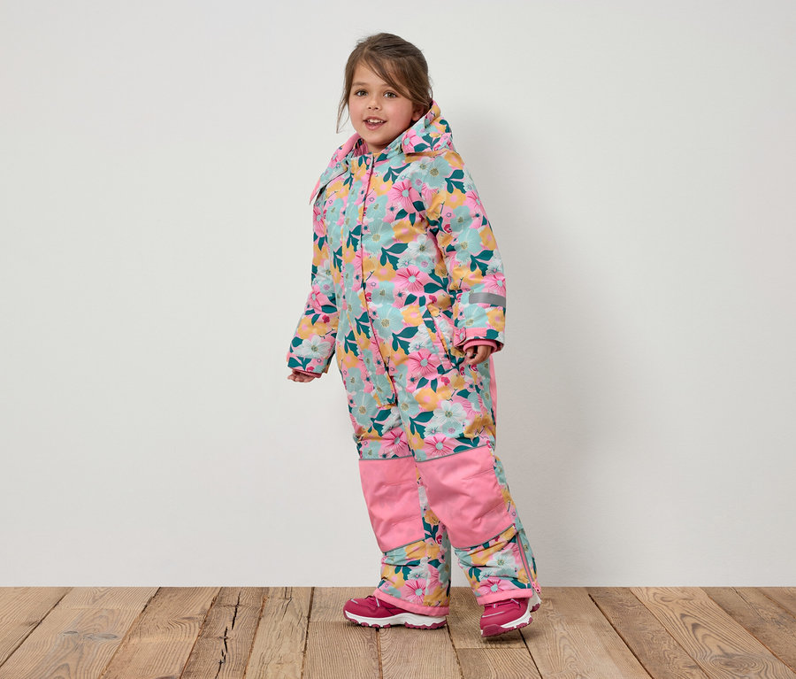 Ein lächelndes Mädchen in einem geblümten Overall mit Kapuze und rosa Schuhen steht auf einem Holzboden.