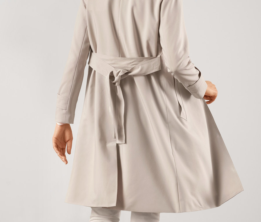 Rückansicht einer Frau, die in einem Trenchcoat, weißen Hosen und Turnschuhen geht.