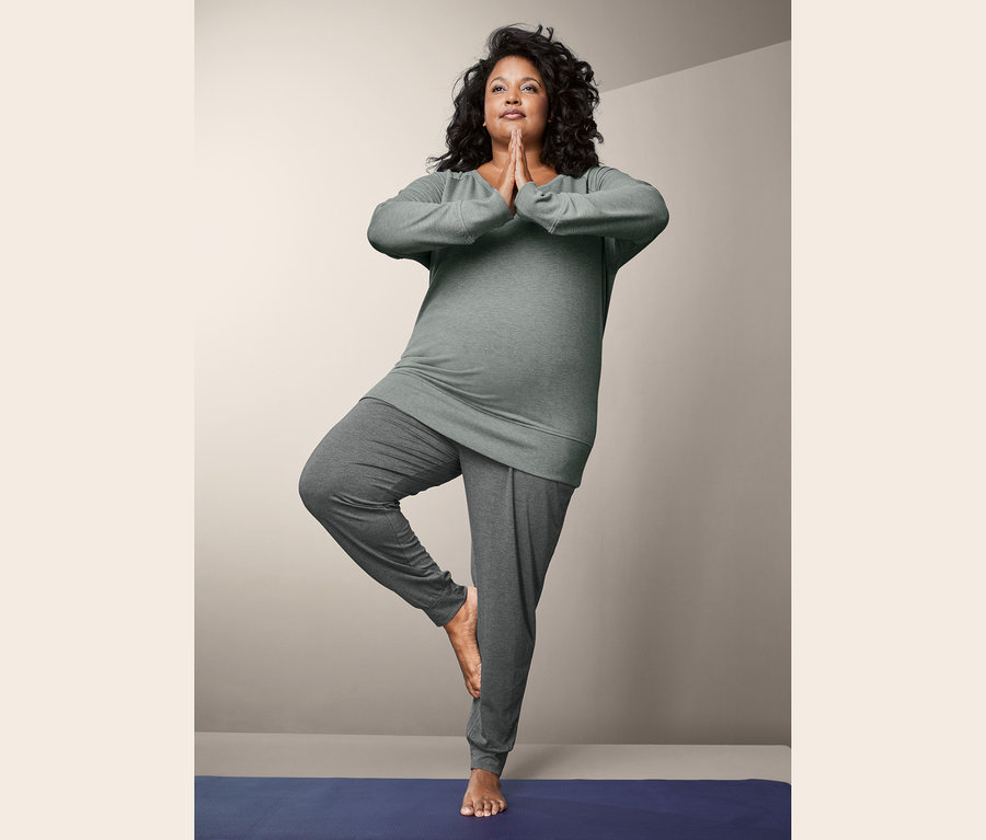 Frau in grauem Yogasweatshirt und Yogahose steht in Baumpose auf blauer Matte.