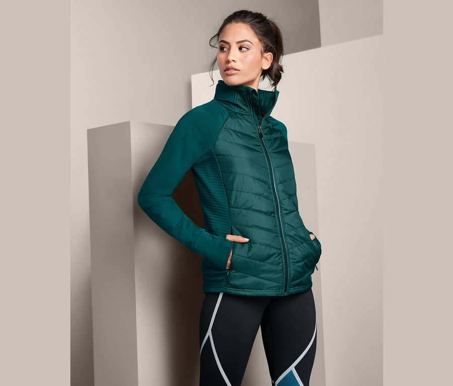 Frau posiert in grüner Thermo-Laufjacke mit verstaubarer Kapuze.