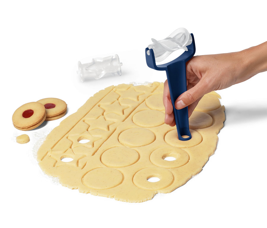Eine Hand benutzt einen Keksroller auf Teig. Daneben liegen zwei gebackene Kekse und ein Aufsatz.