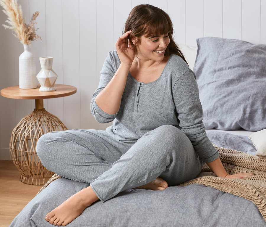 Frau sitzt im grauen Ajour-Pyjama auf dem Bett.