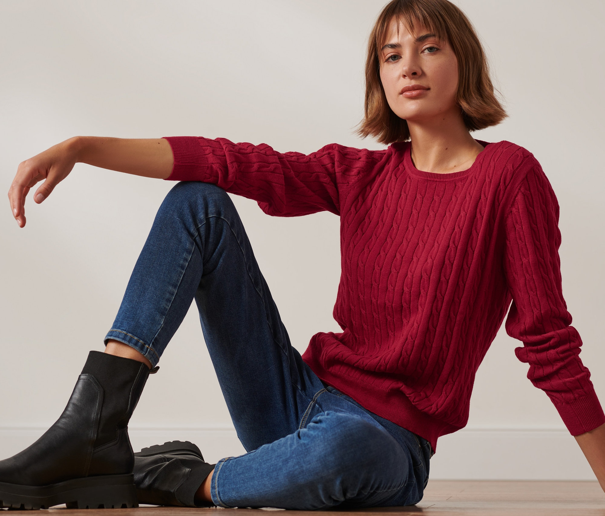 Eine Frau sitzt auf dem Boden und trägt einen roten Zopfstrickpullover, Jeans und schwarze Stiefel.