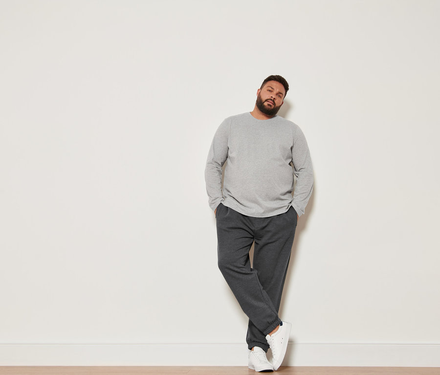 Mann lehnt an Wand, trägt 2 Langarmshirts, Sweathose und Sneaker.