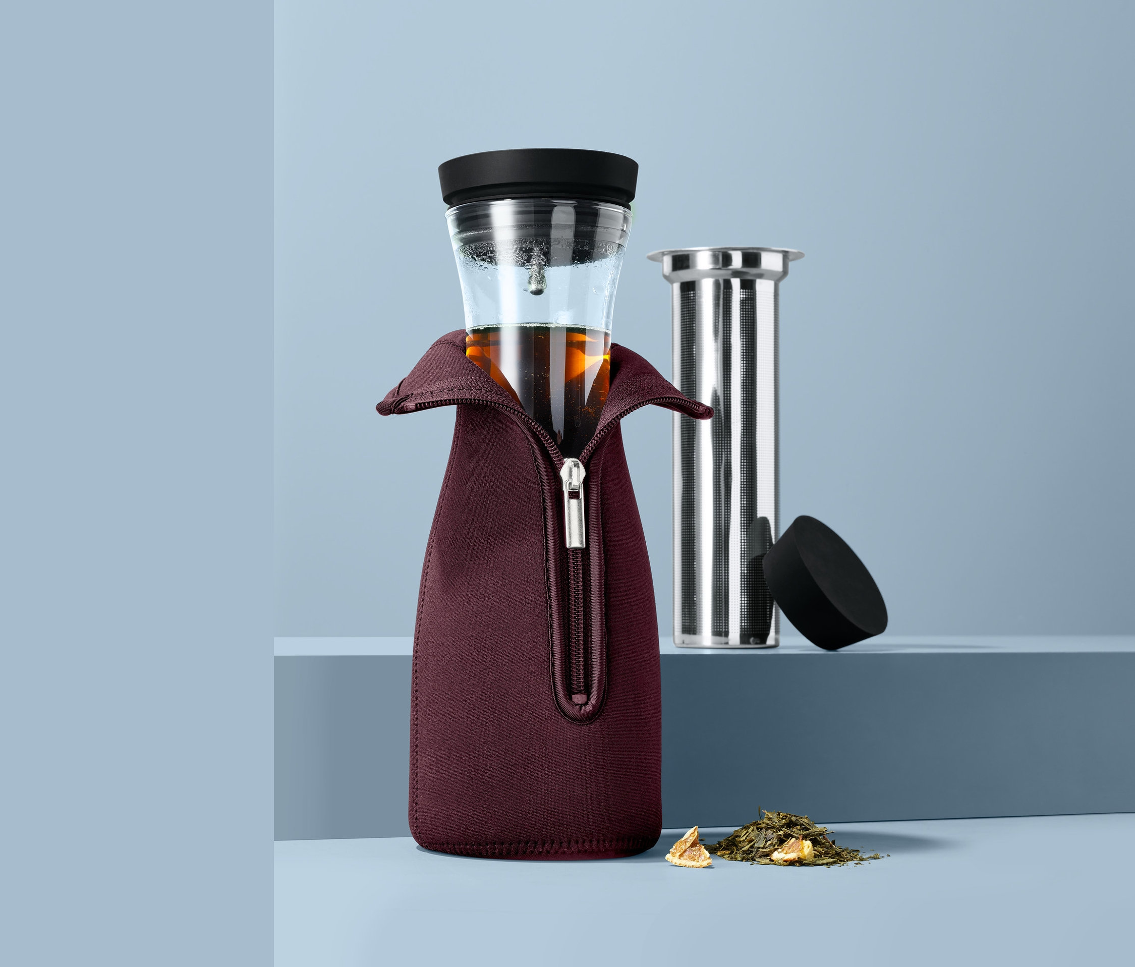 Glas-Teekanne in bordeauxfarbenem Neoprenanzug mit Reissverschluss, daneben Sieb und loser Tee.