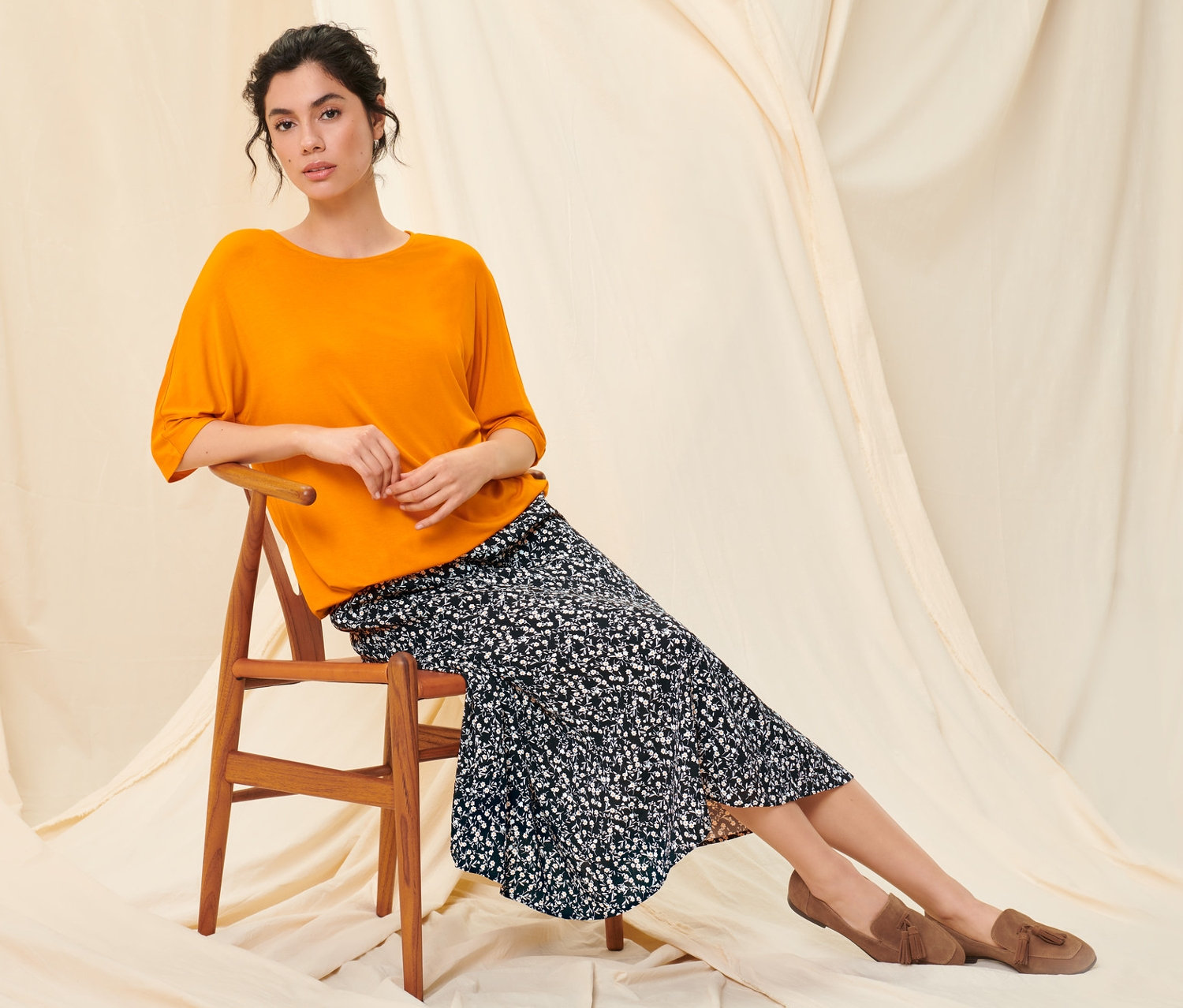 Eine Frau sitzt auf einem Holzstuhl und trägt ein oranges Blusenshirt, einen floralen Midirock und Loafer aus Veloursleder.