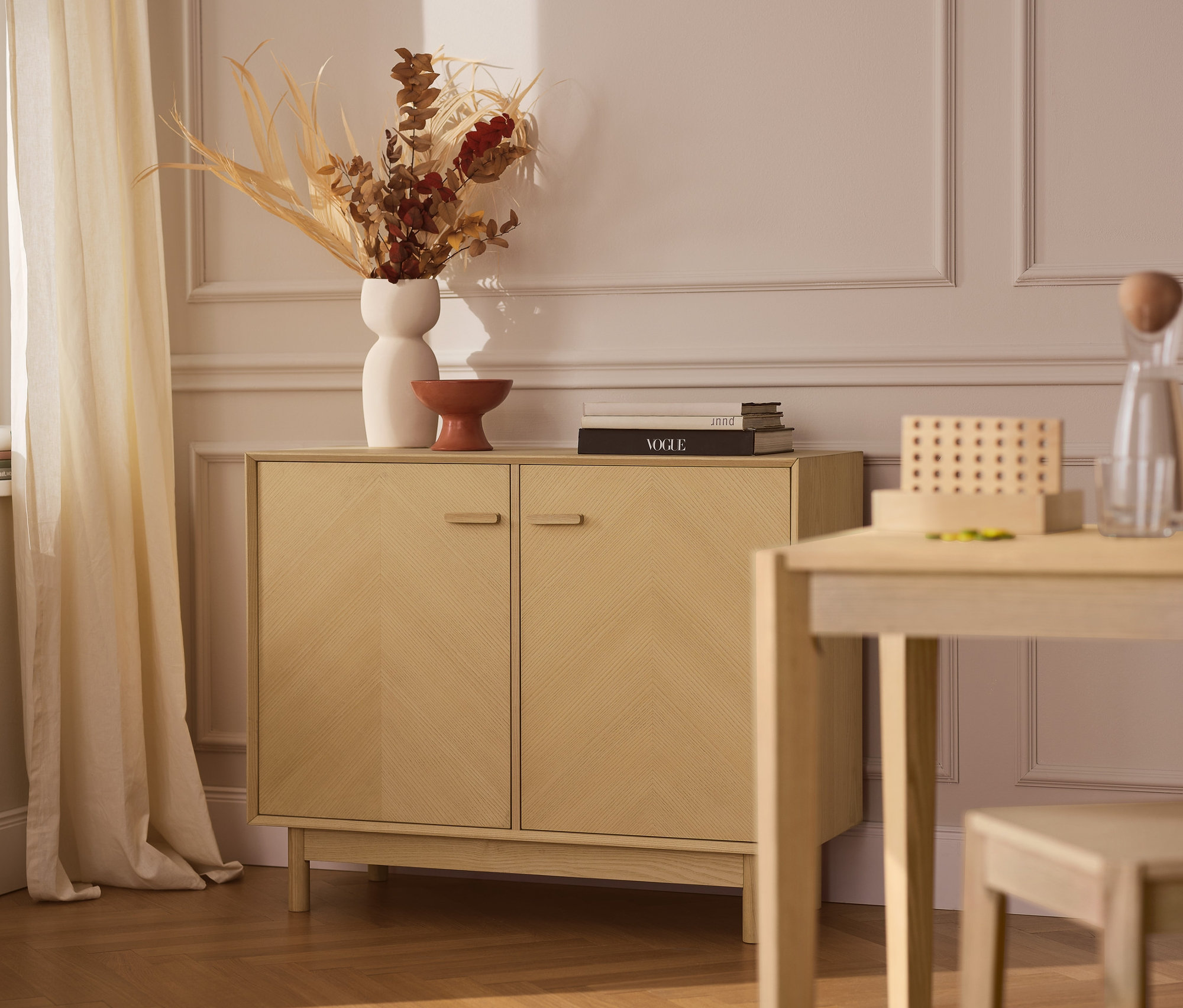 Ein Sideboard aus Eschenholz mit dekorativen Türfronten steht an einer Wand. Auf dem Sideboard stehen eine Vase und Dekorationen. Neben dem Sideboard stehen ein Tisch und ein Stuhl.