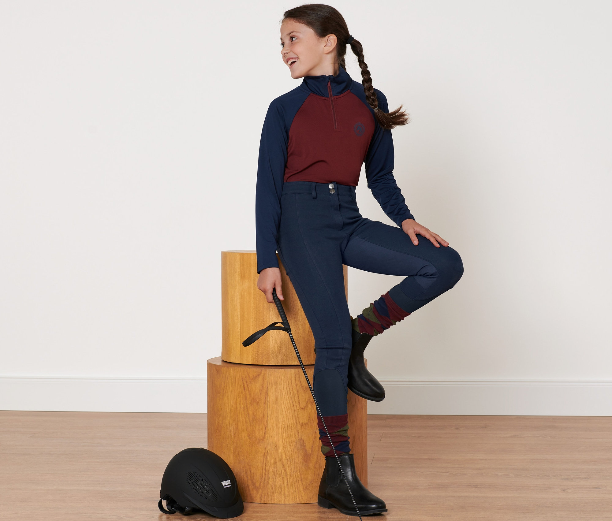 Mädchen sitzt auf Holzblock und trägt Kinder-Vollbesatz-Reithose und Reithelm.