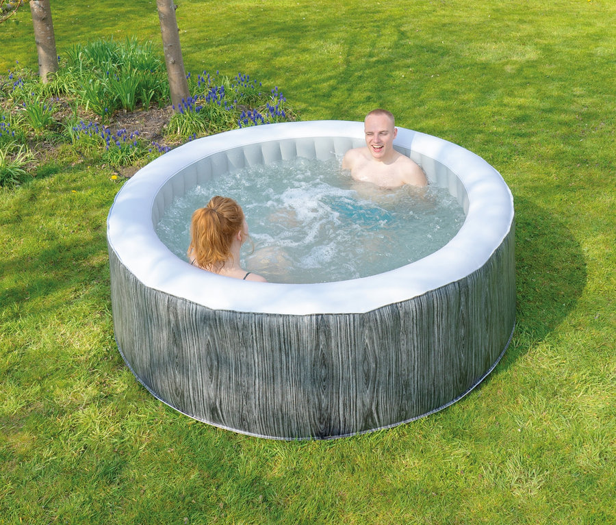 Ein Mann und eine Frau sitzen im EXIT Wood Deluxe Whirlpool für 4 Personen auf einer Rasenfläche vor einem Haus.