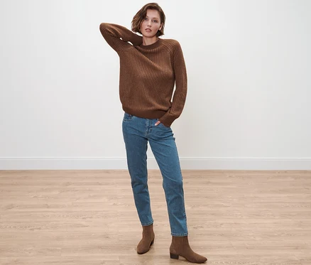 Frau posiert in cognacfarbenem Strickpullover, blauen Jeans und braunen Stiefeletten.