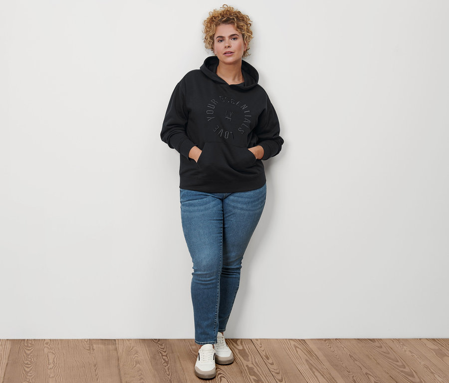 Frau posiert in schwarzem Hoodie, Slim-Denim »Fit Emma« in Midblue Denim und Leder-Sneakern.