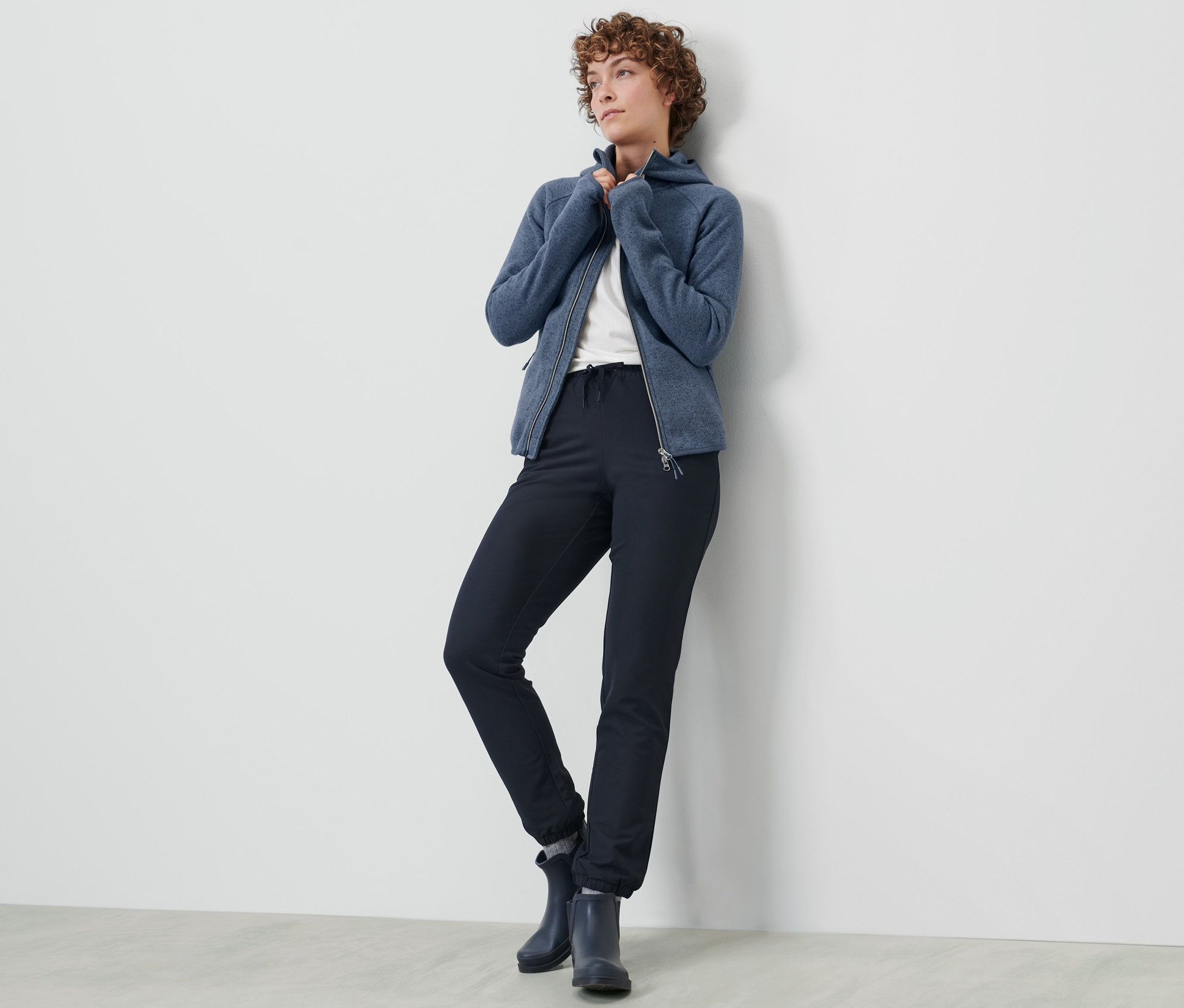 Frau posiert in Softshell-Joggpants und blauer Reißverschlussjacke an einer Wand.