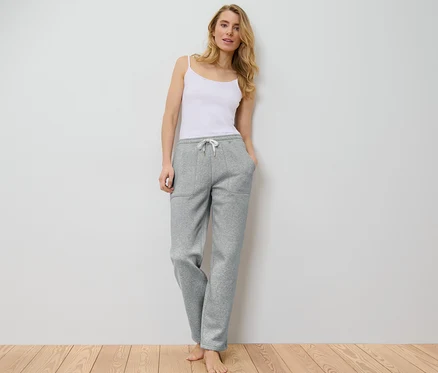 Frau im weißen Top und grauer Loungehose steht an einer weißen Wand.