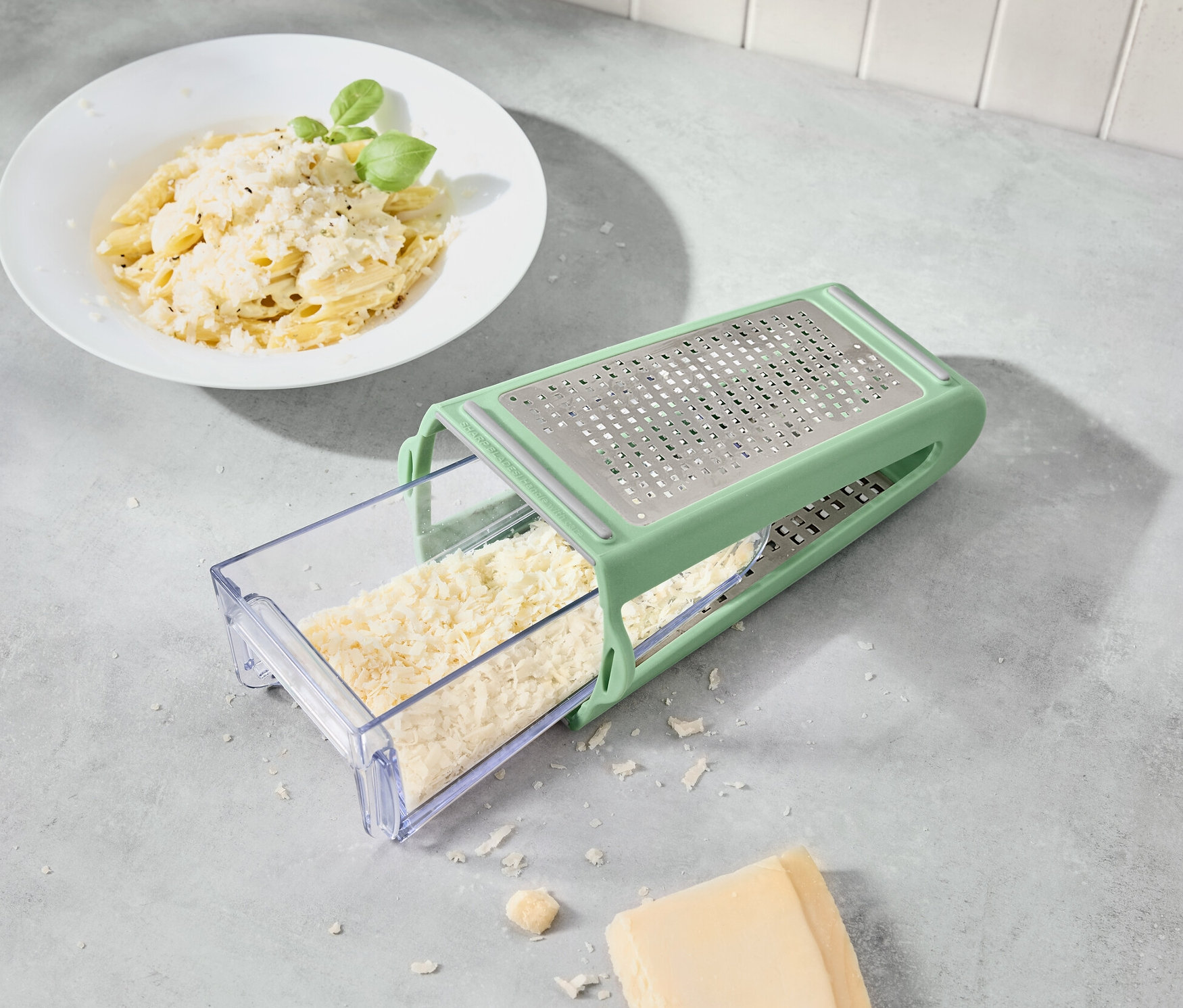 Käse wird mit einer 2-Wege-Reibe mit Auffangbehälter gerieben. Im Hintergrund befindet sich ein Teller mit Nudeln.