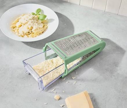 Käse wird mit einer 2-Wege-Reibe mit Auffangbehälter gerieben. Im Hintergrund befindet sich ein Teller mit Nudeln.